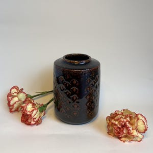 Pode incluir: Um vaso de cerâmica castanho escuro com um padrão de diamante texturizado. O vaso é cilíndrico com uma borda ligeiramente alargada. Dois cravos vermelhos e brancos estão ao lado do vaso. O vaso é provavelmente para decoração de casa.