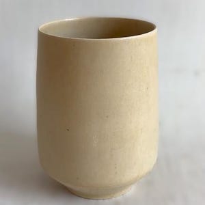 Könnte beinhalten: Becher aus beigefarbener Keramik mit abgerundetem Boden und zylindrischem Körper. Die handgefertigte Keramik hat eine glatte, matte Oberfläche und eine leicht unregelmäßige Form. Die neutrale Farbe und das schlichte Design machen ihn vielseitig einsetzbar.