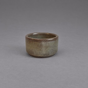 Peut inclure: Un petit bol en céramique cylindrique présentant un émail terreux et tacheté avec des tons mélangés de vert sourd, de brun et de gris. Le récipient possède un bord arrondi lisse et un profil à parois droites, offrant un aspect artisanal.