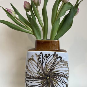 Può includere: Un vaso di ceramica bianco con bordo marrone e motivo floreale, che contiene diversi tulipani rosa con steli e foglie verdi. Il vaso ha una forma rettangolare e un motivo decorativo.