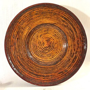 May include: A round, wooden bowl with a spiral design. The bowl is primarily orange and brown, with darker brown concentric circles. The rim is a darker shade of red. The bowl has a glossy finish.