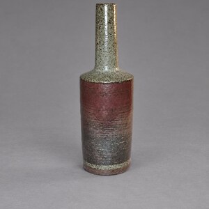 Peut inclure: Une bouteille en céramique haute avec un col étroit et un corps cylindrique. Le col présente un émail gris clair moucheté, tandis que le corps passe d'un brun rougeâtre profond en haut à un gris anthracite foncé à la base avec un bord moucheté.