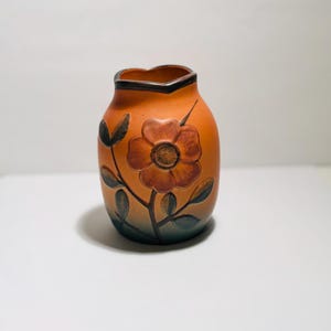 May include: Orange ceramic vase with a floral design. The vase features a dark brown rim and a flower with brown and orange petals. The leaves are green and the background is white.