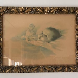 May include: Vintage framed print of a baby. The artwork features a soft, pastel colour palette with a baby lying down. The frame is ornate, with a gold finish and a detailed leaf design. The print is behind glass.