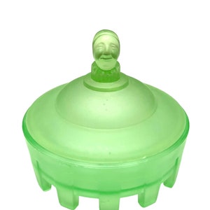 May include: A green glass trinket box with a lid and a smiling face on top. The box is round with a flat base and a lid that fits snugly on top. The glass has a frosted appearance.