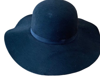 May include: A solid black wide-brimmed hat with a rounded crown and a matching black ribbon band encircling the base. The hat features a soft, felt-like texture and a flexible, wavy brim that curves slightly upward at the edges, creating a classic silhouette.