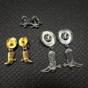 May include: A collection of Western-themed earrings. The earrings include gold-toned cowboy hat and boot dangles, silver-toned hat and boot dangles, and silver-toned bird earrings with turquoise accents. The earrings are displayed on a dark textured surface.