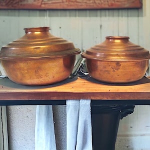 May include: Two copper cooking pots with lids. The pots have a rounded bowl shape with a flared rim and a domed lid. The copper has a warm, reddish-brown color. The pots are sitting on a wooden surface.