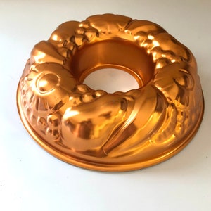 May include: A copper-colored bundt cake pan with a decorative floral and fruit design. The pan has a circular shape with a central hole and a detailed, embossed pattern. The metallic finish reflects light, highlighting the intricate details of the mold.
