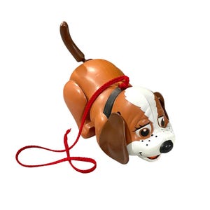 May include: A vintage pull-along toy dog in brown and white with a red leash. The dog has a black collar and a brown tail. The toy is made of plastic and is designed to be pulled along the floor.