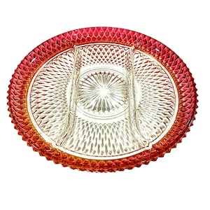 May include: An oval-shaped, divided serving dish with a clear glass center and a red-orange rim. The dish features a diamond-cut pattern throughout the center and a textured, ridged edge. The dish is designed for serving appetizers or snacks.