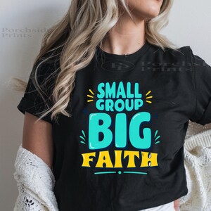 May include: A black short-sleeved t-shirt featuring the text "SMALL GROUP BIG FAITH" printed in a bold, stylized font. The words "SMALL GROUP" are in bright teal, while "BIG FAITH" appears in a larger, vibrant yellow font with teal accents.