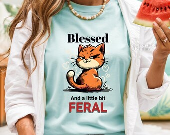 May include: Light blue t-shirt with a cartoon cat illustration. The orange cat has an annoyed expression and is sitting down. The text on the shirt reads "Blessed" above the cat and "And a little bit FERAL" below it.