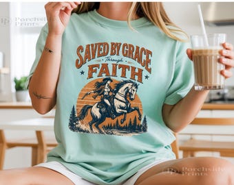 Könnte beinhalten: Ein helles mintgrünes T-Shirt mit einem Grafikdruck, der den Text "SAVED BY GRACE THROUGH FAITH" in orangefarbenen Buchstaben zeigt. Das Design enthält eine Illustration einer Person auf einem Pferd vor einer großen orangefarbenen Sonne und Tannen.