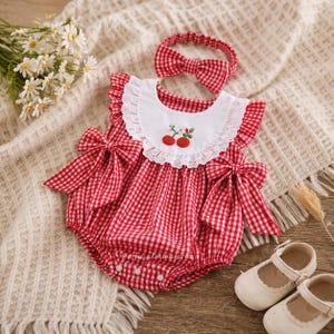 Op de afbeelding: Rood-witte geruite babyromper met een witte kraag met kersenborduurwerk. Inclusief bijpassende haarband met strik. De romper heeft grote strikken op de schouders. Witte babyschoentjes staan ook op de foto.