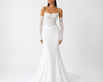May include: A floor-length white lace gown featuring a strapless corset-style bodice with visible boning and matching long, sheer lace sleeves that extend to the wrists. The skirt is fitted through the hips and flares out into a train.