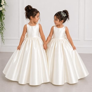 May include: Two floor-length ivory satin dresses featuring a sleeveless bodice, a round neckline, and a full, pleated skirt. Each dress has a defined waistline with a large bow detail at the back, creating a smooth, structured silhouette.