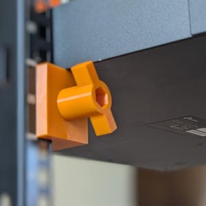 May include: A black network switch mounted on a rack with orange brackets. The switch has multiple ports and is secured with screws. The brackets are a bright orange color and appear to be 3D printed. The rack is black and has vertical rails.