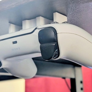 May include: A white PlayStation 5 controller is mounted under a desk using a white holder. The controller has black buttons and triggers. The word "SONY" is printed on the controller.