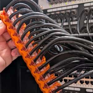 May include: An orange cable organizer holds multiple black network cables. The cables are plugged into a network switch, with additional cables visible in the background. The organizer is held in a person's hand.