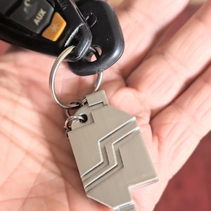 May include: A silver-colored metal keychain with a geometric design. The keychain is attached to a keyring with car keys and a black key fob. The key fob has buttons labeled "AUX" and a star symbol.