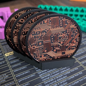 May include: A set of four circular black coasters featuring a raised copper-colored circuit board pattern, held upright in a black plastic stand. The coasters display intricate lines and geometric shapes resembling electronic pathways.