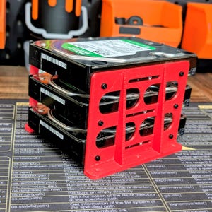 May include: A stack of three silver and black hard drives secured within a bright red, rectangular, 3D-printed plastic storage rack. The rack features a grid-like design with circular cutouts and is fastened with small black screws on the side.