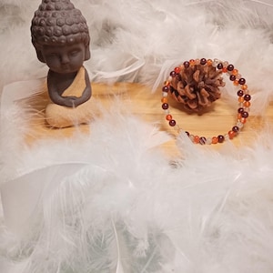 Può includere: Una piccola statua di Buddha marrone con un panno beige è seduta su una pietra chiara. Un braccialetto di perline con perline arancioni e marroni e una pigna sono accanto alla statua. Gli oggetti sono circondati da piume bianche.