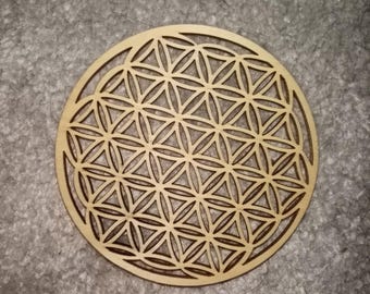 Könnte beinhalten: Ein runder Untersetzer aus Holz mit einem lasergeschnittenen Flower-of-Life-Design. Der Untersetzer besteht aus hellfarbenem Holz und weist ein sich wiederholendes geometrisches Muster aus sich überlappenden Kreisen und Dreiecken auf. Der Untersetzer hat einen Durchmesser von etwa 10 cm.