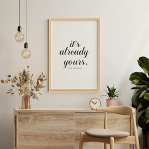 Könnte beinhalten: Gerahmter Druck mit dem Text "it's already yours." in minimalistischem Stil. Der Druck wird über einem Holzschrank mit einer Vase mit Trockenblumen, einer kleinen Uhr und einem Stuhl angezeigt. Zwei hängende Glühbirnen sind sichtbar.