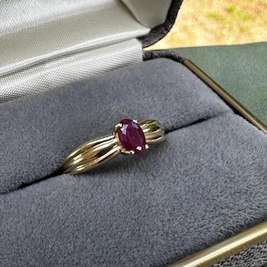 May include: A gold ring with a ruby gemstone, presented in a grey velvet jewellery box. The oval-shaped ruby is a deep red. The ring features a distinctive, flowing design.