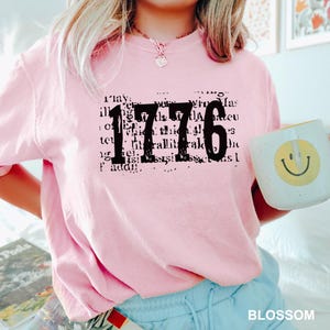 May include: A light pink short-sleeve t-shirt featuring the black text 1776 printed over a background of faint, illegible black script. The shirt has a crew neckline and a relaxed fit, showcasing a casual style with a soft, solid-colored fabric.