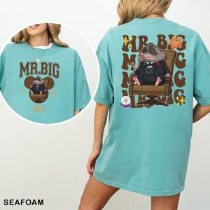A woman wearing a teal t-shirt with a cartoon image of a man wearing a suit and tie, along with the text "MR. BIG" and "AIR MUGS" printed on the back.