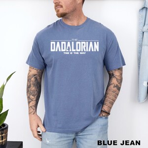 A man wearing a blue t-shirt with the text "THE DADALORIAN" printed on it, standing against a white wall.