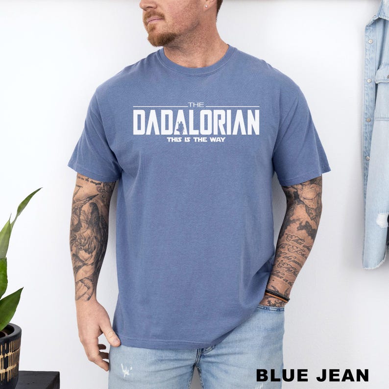 A man wearing a blue t-shirt with the text "THE DADALORIAN" printed on it, standing against a white wall.