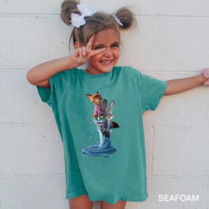 A young girl wearing a green t-shirt with a cartoon character printed on it, smiling and making a peace sign gesture.