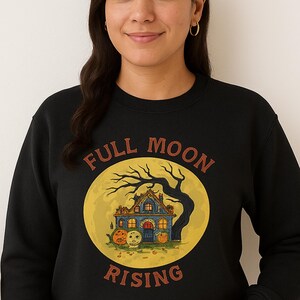 May include: A black long-sleeve crewneck sweatshirt featuring a graphic print of a house with pumpkins and a bare tree set against a large yellow moon. The text "FULL MOON RISING" is printed in a curved, rust-colored font around the circular design.