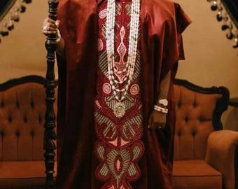 Puede incluir: Un atuendo tradicional granate con intrincados bordados y abalorios. El conjunto incluye un sombrero a juego y un largo collar de cuentas. La persona sostiene un bastón negro. El logotipo de CJ Fashion Demos está en la esquina superior derecha.