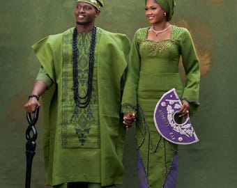 Puede incluir: Una pareja con atuendos tradicionales verde oliva a juego. El hombre lleva una túnica y pantalones con un collar de cuentas negras y sostiene un bastón tallado. La mujer lleva un vestido y un pañuelo en la cabeza a juego, sosteniendo un abanico morado con texto.