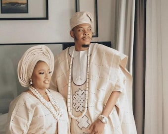 May include: A couple in traditional Nigerian attire. The man wears a cream-colored outfit with intricate embroidery and a matching cap. The woman wears a cream-colored head wrap and matching dress, adorned with beaded jewelry and a fan.