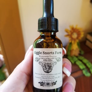 Puede incluir: Una peque&ntilde;a botella de vidrio marr&oacute;n con un gotero negro. La botella est&aacute; etiquetada con "Giggle Snorts Farm Wildcrafted Tincture Wild Violet". La etiqueta tambi&eacute;n incluye instrucciones de uso e ingredientes: "Place 1 dropperful under tongue or in glass of water. Ingredients: Organic Cane Alcohol, Wild Violets & Pure Love!"
