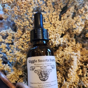 May include: A glass bottle with a dropper top, labeled "Giggle Snorts Farm Wildcrafted Tincture Yarrow". The bottle is sitting in a basket of dried flowers.