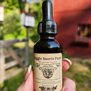 May include: A small glass bottle with a dropper top, labeled "Giggle Snorts Farm Wildcrafted Tincture Lemon Balm". The label has a floral design and the text "Directions: Place 1 dropperful under tongue or in glass of water. Ingredients: Organic Cane Alcohol, Lemon Balm & Pure Love!"