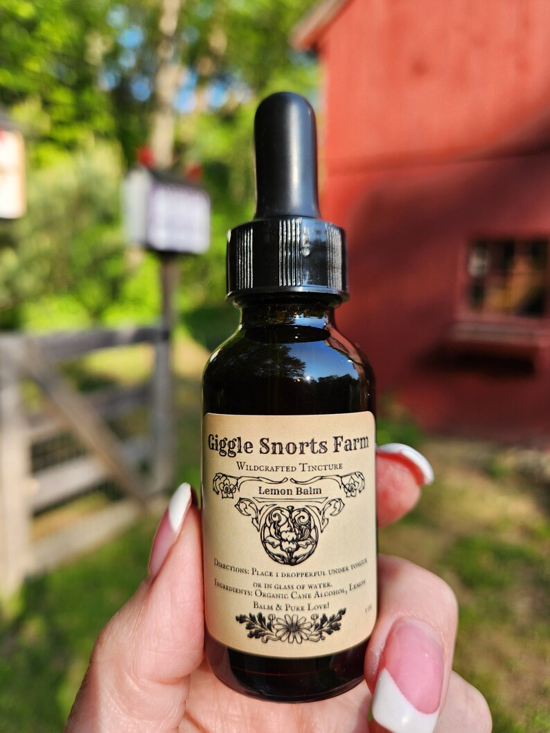 May include: A small glass bottle with a dropper top, labeled "Giggle Snorts Farm Wildcrafted Tincture Lemon Balm". The label has a floral design and the text "Directions: Place 1 dropperful under tongue or in glass of water. Ingredients: Organic Cane Alcohol, Lemon Balm & Pure Love!"