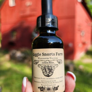 May include: A glass bottle with a dropper top, labeled "Giggle Snorts Farm Wildcrafted Tincture Lemon Balm". The label has a floral design and the text "Directions: Place 1 dropperful under tongue or in glass of water. Ingredients: Organic Cane Alcohol, Lemon Balm & Pure Love!"