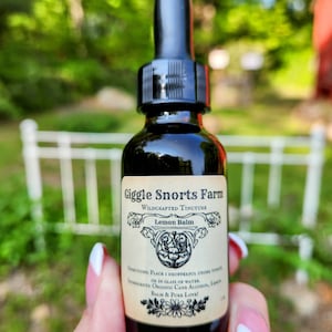 May include: A small glass bottle with a dropper top, labeled "Giggle Snorts Farm Wildcrafted Tincture Lemon Balm". The label has a floral design and the text "Directions: Place 1 dropperful under tongue or in glass of water. Ingredients: Organic Cane Alcohol, Lemon Balm & Pure Love!"