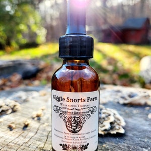 May include: A brown glass bottle with a dropper top and a white label that reads "Giggle Snorts Farm Wildcrafted Tincture Japanese Knotweed". The label has a decorative border and a small illustration of a flower.