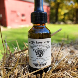 May include: A brown glass bottle with a black dropper top and a white label that reads "Giggle Snorts Farm Wildcrafted Tincture Yarrow". The bottle is sitting on a bed of straw.