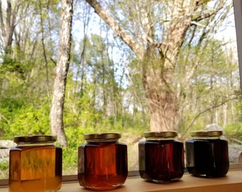 May include: Four glass jars with gold lids filled with different shades of honey. The jars are lined up on a wooden surface in front of a window with a view of a forest.
