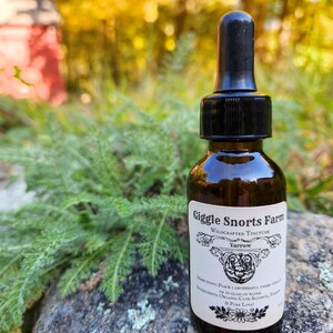 May include: A small brown glass bottle with a black dropper top. The bottle is labeled "Giggle Snorts Farm Wildcrafted Tincture Yarrow". The label also includes directions for use and ingredients: "Place 1 dropperful under tongue or in glass of water. Ingredients: Organic Cane Alcohol, Yarrow & Pure Love!"